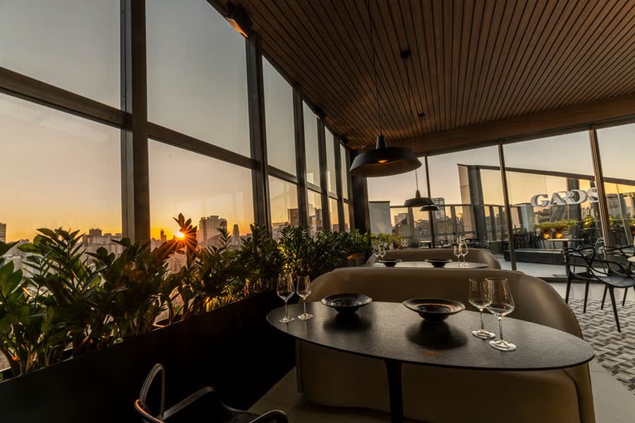 Mesa posta junto às janelas, com o pôr do sol atrás das plantas